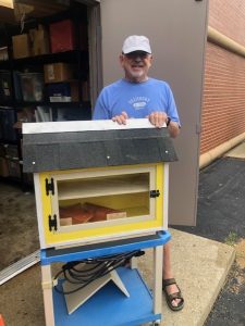 Man in baseball cap and blue t-shirt stands with a yellow Little Free Library