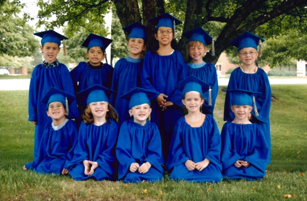 Eleven young children in blue graduation caps and gowns sitting under a tree