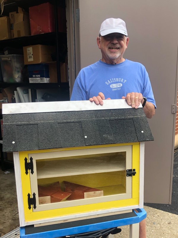 Man in blue shirt and white hat standing next to a yellow Little Free Library
