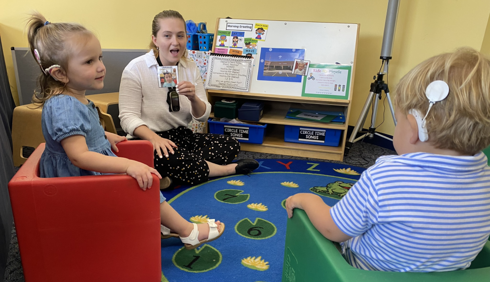 Woman talks to two children seated in chairs wearing cochlear implants