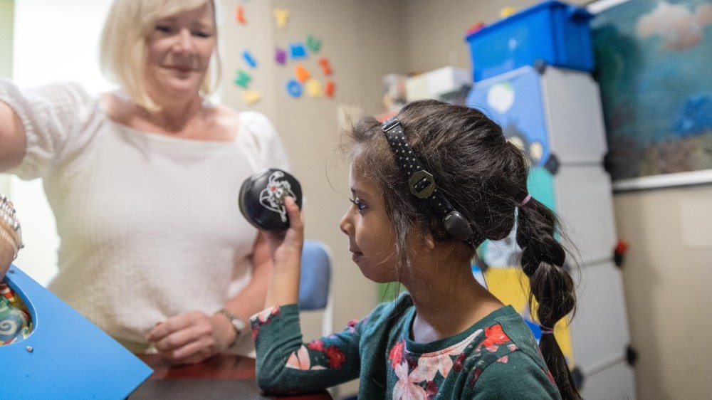 A young girl participates in an audiology test while a female audiologist looks on