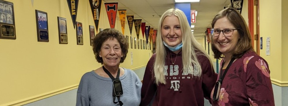 A woman in grey, a woman in maroon, and a womain in a flowerd blouse smile in a hallway lined with college pennants.