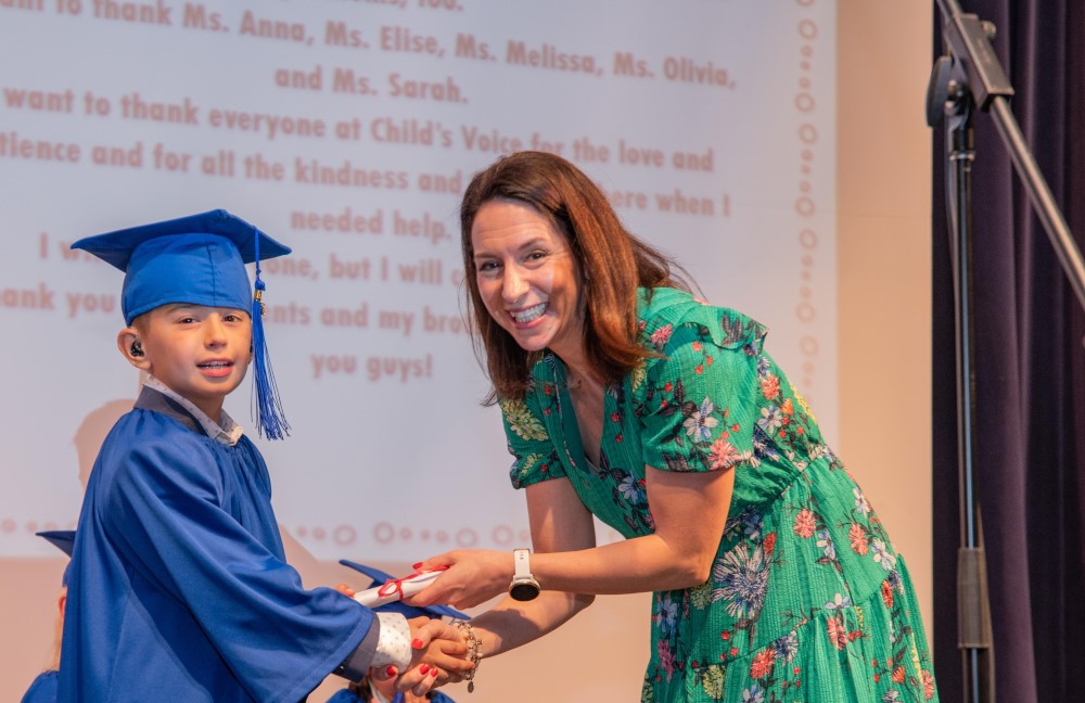 A young boy in a graduation cap and gown takes a diploma from a woman in a green dress