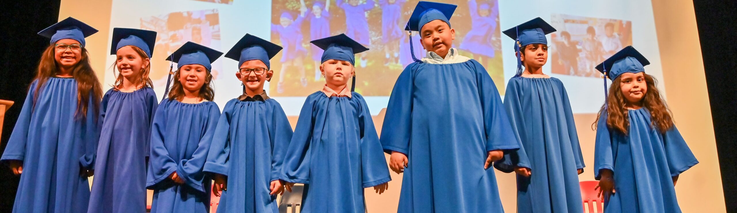 Eight young children in blue graduation gowns stand on stage