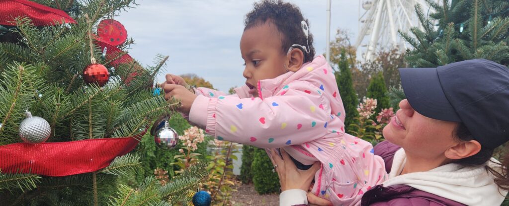 A woman in a baseball cap helps a young girl wearing a cochlear implant hang an ornament on a holiday tree