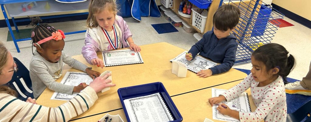 A group of young students sits around a table while a teacher directs their work.