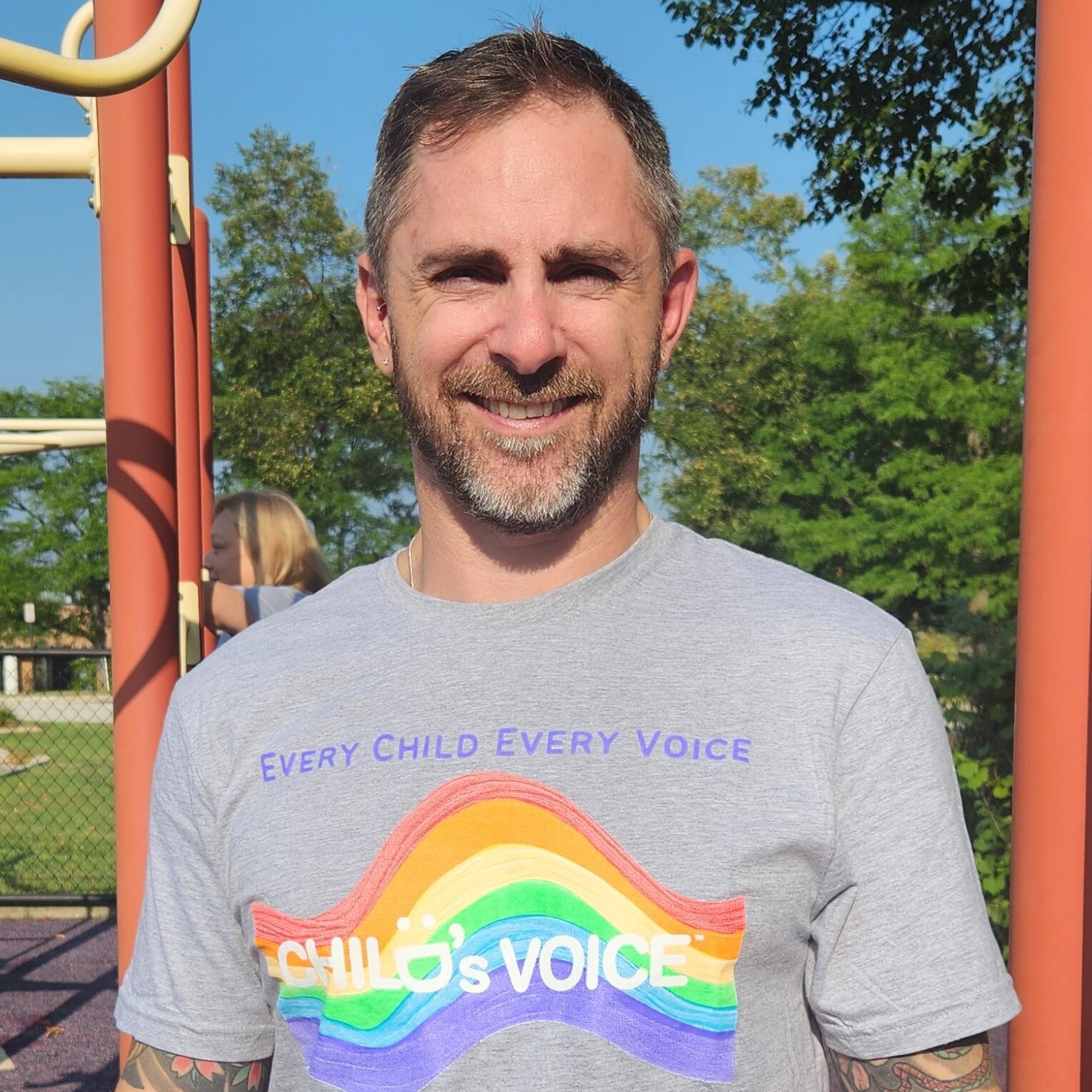 A man in a grey t-shirt with a rainbow on front stands in front of a playground